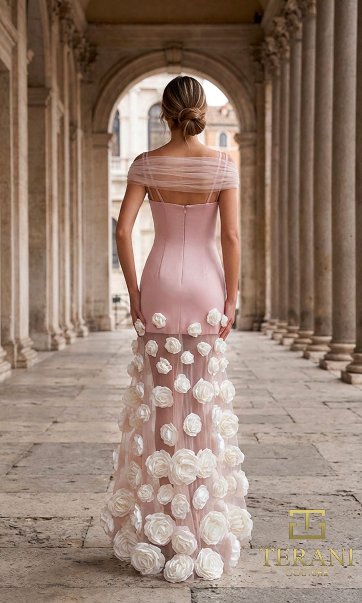 A woman stands in a stone-columned walkway, photographed from behind, wearing the Long Formal Dress: Terani 261P6098—a light pink evening gown with sheer shoulders and floral appliques on the skirt.